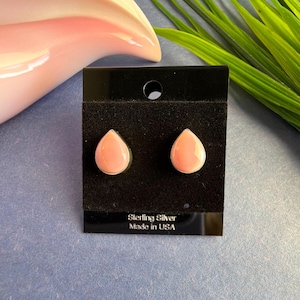 May include: A pair of teardrop-shaped stud earrings with a light pink center and a silver border, displayed on a black card. The card reads "Sterling Silver Made in USA". The earrings are set against a blue background.