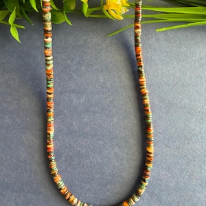 May include: A necklace composed of small, flat, circular disc beads in a variety of colors including turquoise, orange, brown, and cream. The beads are strung together in a repeating pattern, creating a multi-colored, textured strand of jewelry.