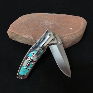 May include: A silver folding knife with a partially open, polished steel blade. The handle features turquoise, red, black, and white inlay designs. The knife is set against a dark background and a reddish-brown stone.