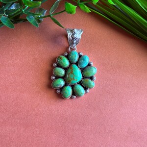 May include: A silver pendant with a central turquoise stone surrounded by oval-shaped turquoise stones. The pendant has a detailed silver bail and is set against a peach background. Green foliage is visible in the background.