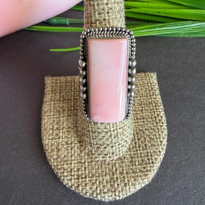May include: A silver ring featuring a rectangular, pale pink stone. The stone is set within a decorative frame with a beaded edge. The ring is presented on a small, beige fabric stand, highlighting the jewellery's design and colour.