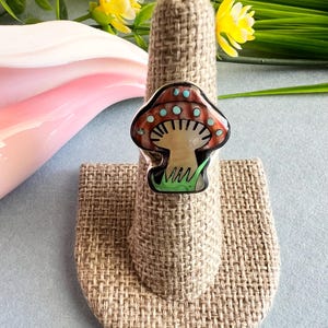 Puede incluir: Un anillo de plata con un diseño de hongo hecho de piedras rojas, marrones, blancas y verdes. El hongo tiene puntos blancos en la parte superior.