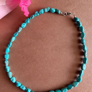 May include: A turquoise stone necklace with a silver clasp. The necklace is made of small, irregularly shaped turquoise beads. The beads are a vibrant blue-green color. The necklace is displayed on a brown surface with a pink flower in the background.