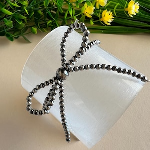 May include: A silver beaded hair tie with a bow design. The hair tie is tied around a white ceramic vase.