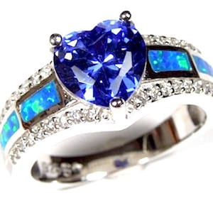 Op de afbeelding: Een zilveren ring met een grote blauwe hartvormige edelsteen omringd door kleinere heldere edelstenen. De band is versierd met blauwe en groene opaal accenten.