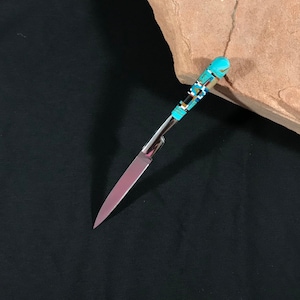 May include: A decorative knife with a polished steel blade and a handle inlaid with turquoise, black, white, and blue stones. The knife is set against a black background and a tan rock.
