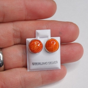 May include: A pair of round stud earrings featuring vibrant orange, marbled cabochons set in polished metal. The earrings are mounted on a small white display card with the text STERLING SILVER printed in black at the bottom of the card.