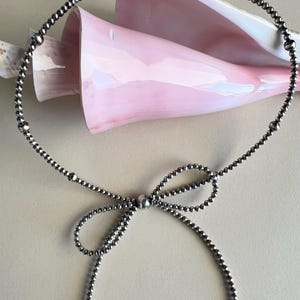 May include: A silver beaded necklace with a bow tied in the center. The necklace is lying on a light beige surface.