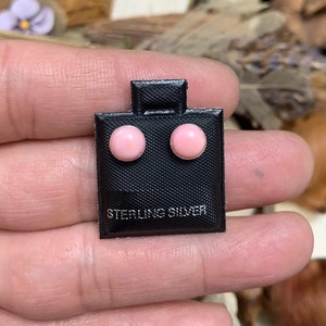 May include: A pair of round, light pink stud earrings set in sterling silver. The earrings are mounted on a small, black, textured rectangular display card with the text STERLING SILVER printed in white at the bottom of the card.