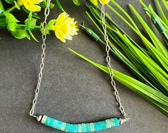 May include: A silver chain necklace with a row of turquoise-colored rectangular beads. The necklace is displayed against a backdrop of yellow flowers and green foliage, creating a vibrant contrast.