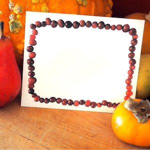 May include: A seasonal still life composition with a red pear, orange persimmon, and pumpkins. A white card, framed with cranberries, is centrally placed. The arrangement is set on a wooden surface, suggesting an autumnal theme.