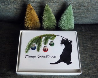 May include: A box of Christmas cards featuring a black cat silhouette hanging from a decorated branch with ornaments. The words "Merry Christmas" are printed below the image. Three small, decorative Christmas trees are in the background.