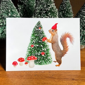 May include: A Christmas card featuring a squirrel wearing a red Santa hat, holding a red and white mushroom. The squirrel is standing in front of a green Christmas tree decorated with red and white mushrooms.