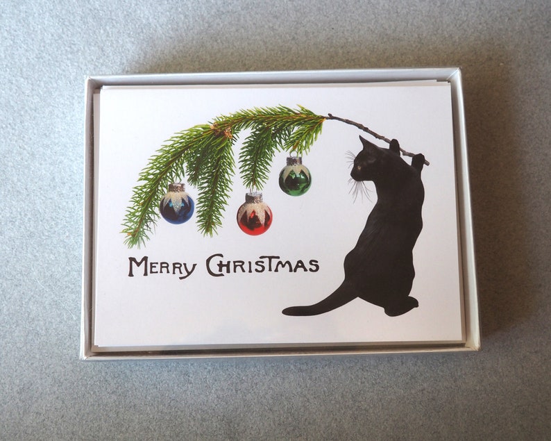 Op de afbeelding: Set kerstkaarten met een zwarte kat die een tak met ballen vasthoudt. De tak heeft groene naalden en kleurrijke ballen. De woorden "MERRY CHRISTMAS" staan eronder gedrukt.
