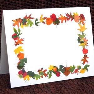 May include: A white card with a border of fall leaves in shades of red, orange, yellow, and brown. The leaves are arranged in a circle around the edge of the card, leaving a blank space in the center.