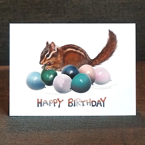 May include: A birthday card featuring a cartoon chipmunk holding a green balloon surrounded by colorful balloons with the text "Happy Birthday".