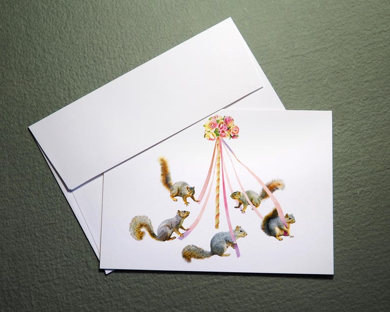 May include: A white card with a maypole decorated with pink and white flowers. Five gray squirrels are climbing the maypole, each holding a pink ribbon.