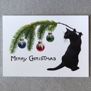 Op de afbeelding: Een witte kerstkaart met een zwarte katten silhouet die een tak vasthoudt met groene dennennaalden en kerstballen. De woorden "MERRY CHRISTMAS" staan in zwarte letters eronder. De ballen zijn blauw, rood en groen.