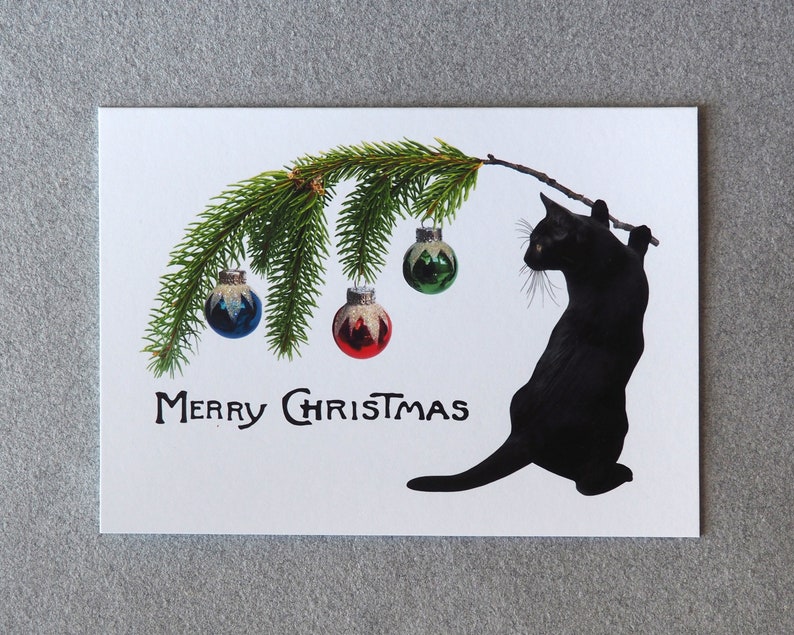 Op de afbeelding: Een witte kerstkaart met een zwarte katten silhouet die een tak vasthoudt met groene dennennaalden en kerstballen. De woorden "MERRY CHRISTMAS" staan in zwarte letters eronder. De ballen zijn blauw, rood en groen.