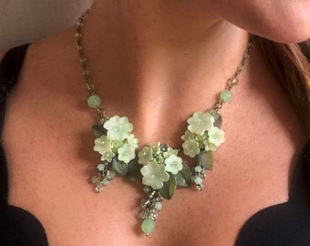 May include: A necklace featuring pale green floral charms and dark green leaf accents arranged in a cluster. The piece includes small green beads and a metallic chain, with a central floral drop element hanging from the main decorative section.
