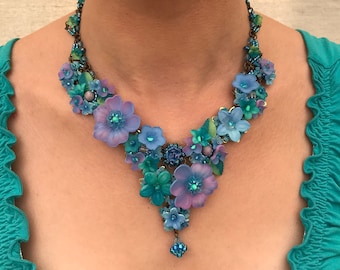 Può includere: Una collana importante caratterizzata da una serie di elementi floreali blu, viola e turchesi. Il design incorpora fiori di varie dimensioni, piccole perline ed elementi metallici disposti a V, con un singolo ciondolo di perline al centro.