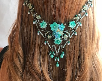 May include: A decorative headpiece with a floral design. The headpiece features turquoise and light blue flowers, white and turquoise beads, and dangling turquoise beads. The metal is a dark bronze color. The headpiece is worn on the head.