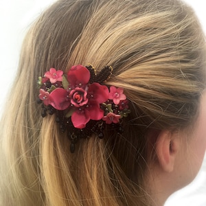 以下が含まれることがあります： 黒ビーズと濃い緑色の茎が付いたピンクの花のヘアクリップ。クリップは女性のブロンドの髪に留められています。