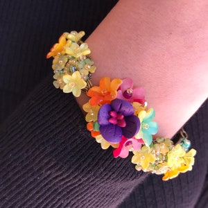 May include: A floral bracelet featuring a cluster of colorful flowers in shades of yellow, orange, pink, purple, and blue. The bracelet is adorned with small, sparkling beads and is worn on a wrist against a black background.
