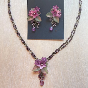 Pu&ograve; includere: Un set di collana e orecchini con motivi floreali. La collana ha una catena con un ciondolo di fiori viola e perline pendenti. Gli orecchini sono a clip con grappoli floreali abbinati. Il set &egrave; esposto su una superficie di legno chiaro.
