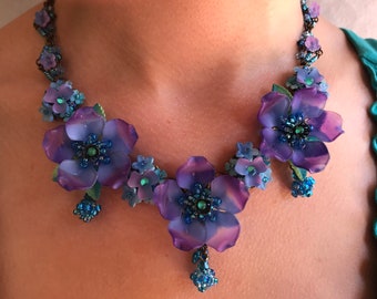 May include: A purple and blue floral necklace with a beaded design. The necklace features large, layered flowers with a beaded center and smaller flowers interspersed throughout.