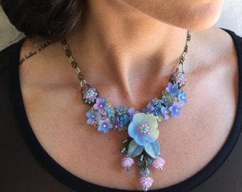 Può includere: Un delicato set di collana e orecchini floreali con fiori blu, viola e rosa. La collana presenta un design a cascata con un grande fiore blu al centro.