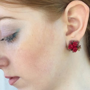 May include: A woman wearing a red and green beaded necklace and red flower earrings.