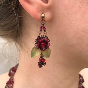 Könnte beinhalten: Ohrringe mit Anhänger mit einer dunkelroten Rose, goldfarbenen Blättern und roten Perlen. Die Ohrringe sind an einem kleinen goldfarbenen Stecker befestigt. Die Ohrringe sind Teil eines Schmucksets.