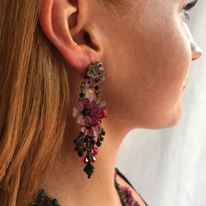 Op de afbeelding: Een paar lange, hangende oorbellen met een cluster van roze, rode en groene bloemen en zwarte kralen.