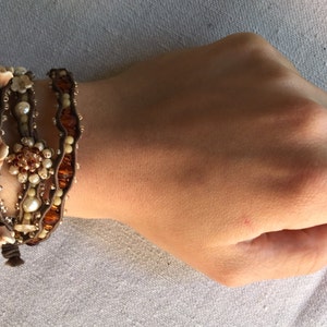 May include: A multi-strand wrap bracelet featuring brown leather cords adorned with cream-colored pearls, amber-toned beads, and decorative floral accents. The bracelet is wrapped around a wrist, showcasing its intricate design.