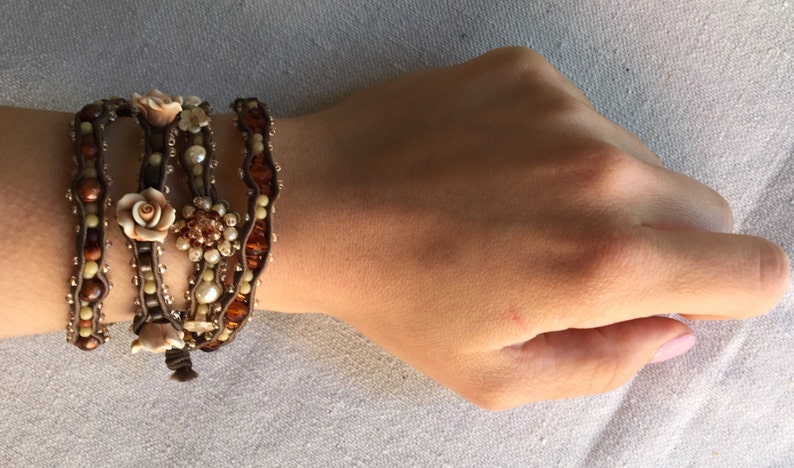 May include: A multi-strand wrap bracelet featuring brown leather cords adorned with cream-colored pearls, amber-toned beads, and decorative floral accents. The bracelet is wrapped around a wrist, showcasing its intricate design.