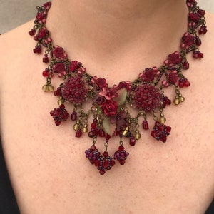 Peut inclure: Un collier floral bordeaux avec des perles complexes et des accents pendants. Le collier présente des grappes de petites fleurs et perles rouges, avec une chaîne et des accents en métal doré. Le design comprend un arrangement floral central avec des accents roses et verts.