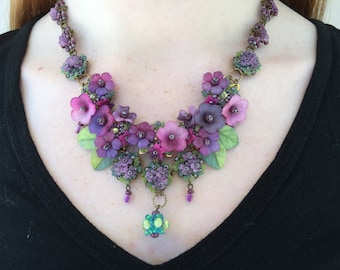 May include: A floral statement necklace featuring clusters of purple and magenta flowers, green leaves, and small beads. The design includes a central dangling bead element in shades of blue and green, all connected by a bronze-toned metal chain.