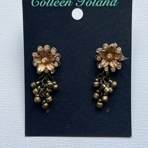 May include: Gold-toned earrings with a flower design and dangling beads, displayed on a black card. The earrings feature clear crystals and are labelled with the text "Colleen Toland".