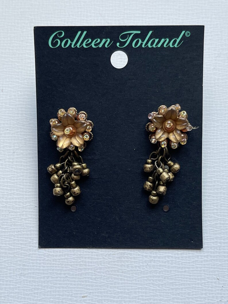 May include: Gold-toned earrings with a flower design and dangling beads, displayed on a black card. The earrings feature clear crystals and are labelled with the text "Colleen Toland".