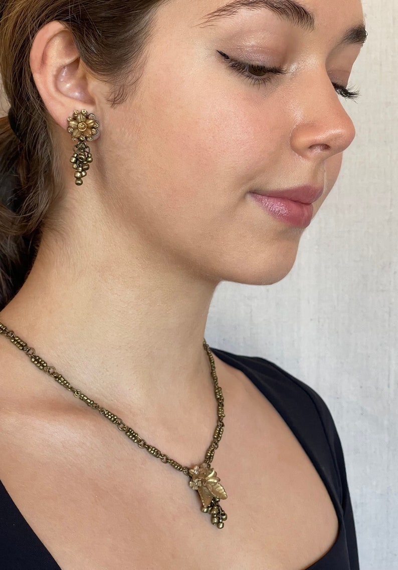 May include: A gold-toned jewellery set featuring a floral design. The set includes a necklace with a pendant and matching earrings. The earrings and pendant have a cluster of small beads. The necklace chain is made of small, connected beads.