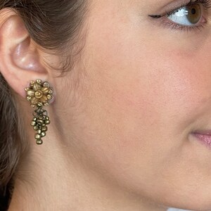 May include: A vintage-style gold-toned earring with a floral design and dangling bead accents. The earring features a flower-shaped stud with small, sparkling embellishments around the petals. Below the flower, a cluster of small beads hangs.