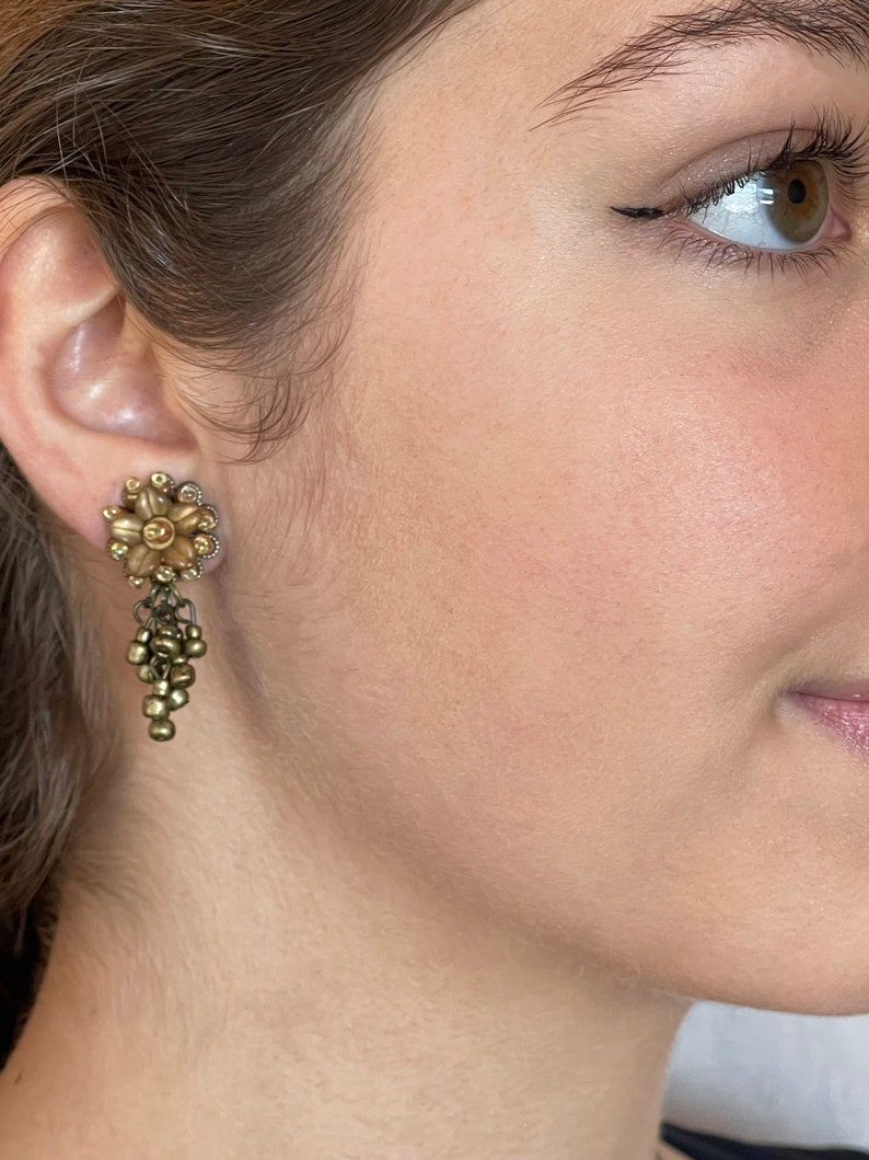 May include: A vintage-style gold-toned earring with a floral design and dangling bead accents. The earring features a flower-shaped stud with small, sparkling embellishments around the petals. Below the flower, a cluster of small beads hangs.