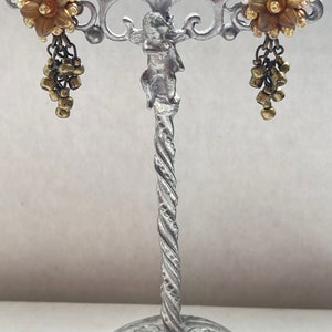 May include: A pair of vintage earrings on a silver stand. Each earring has a gold-toned flower with clear rhinestones and dangling gold beads. The stand features a cherub and a twisted stem. The earrings are a classic design.