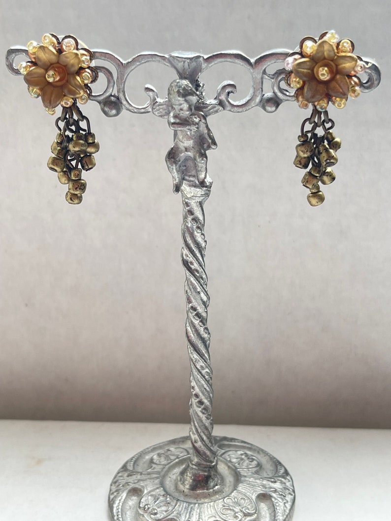 May include: A pair of vintage earrings on a silver stand. Each earring has a gold-toned flower with clear rhinestones and dangling gold beads. The stand features a cherub and a twisted stem. The earrings are a classic design.