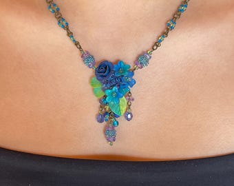 May include: A blue and green floral necklace with a cluster of blue flowers and green leaves. The necklace is made of metal and beads.