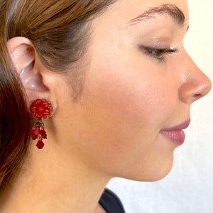 May include: Red flower-shaped dangle earrings with red and clear crystal beads. The earrings are worn by a person with brown hair.