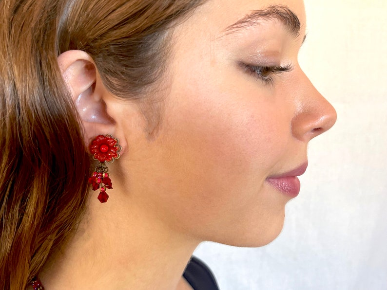 May include: Red flower-shaped dangle earrings with red and clear crystal beads. The earrings are worn by a person with brown hair.