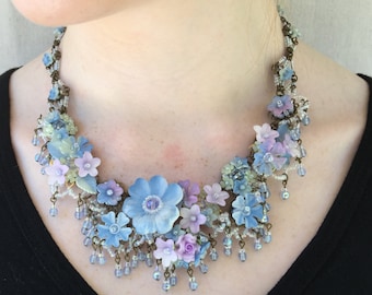 Può includere: Una delicata collana con una cascata di fiori blu, viola e verdi. La collana è realizzata con perline e ha uno stile vintage.