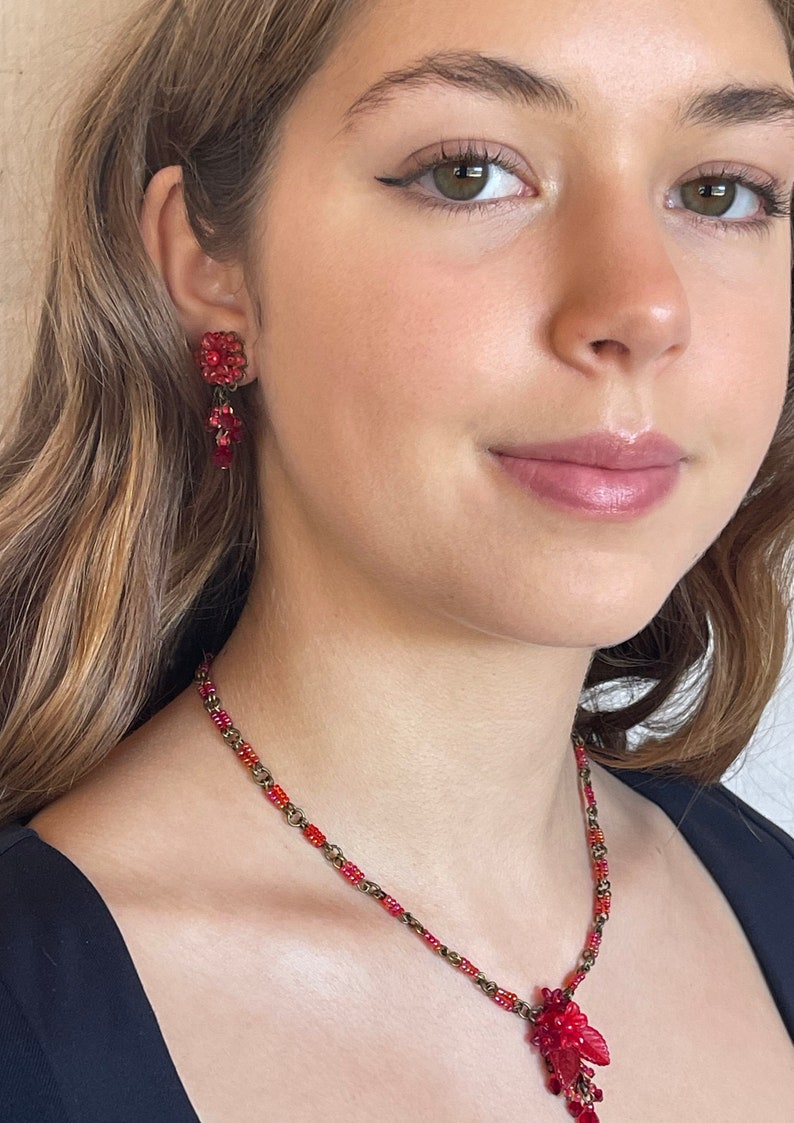 May include: A red beaded necklace with a floral pendant and matching earrings. The necklace and earrings are made of small red beads and have a vintage look.